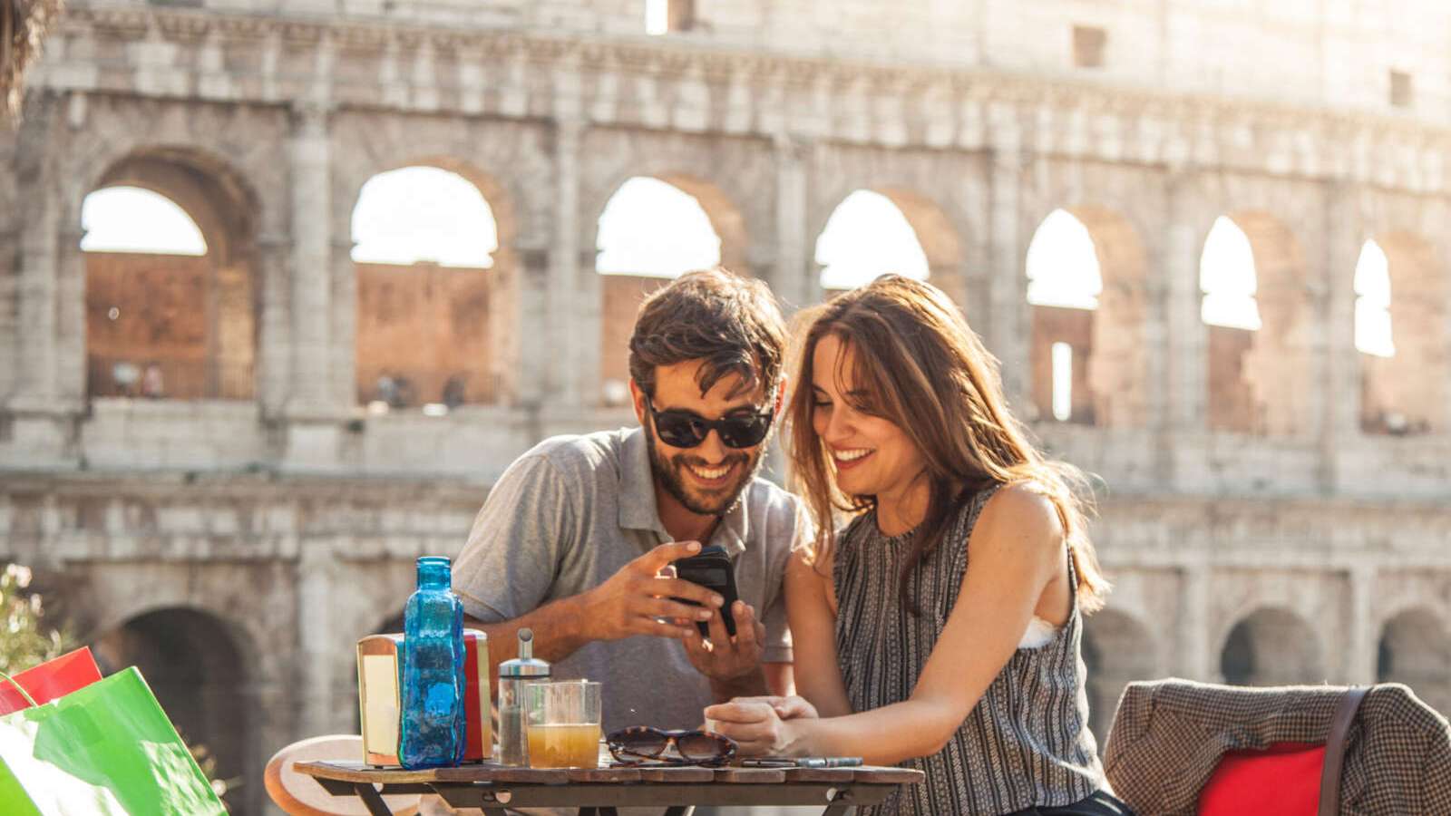 Happy young couple tourists using smartphone sitting at bar restaurant in front of colosseum in rome at sunset with coffee shopping bags smiling having fun texting browsing and sharing pictures